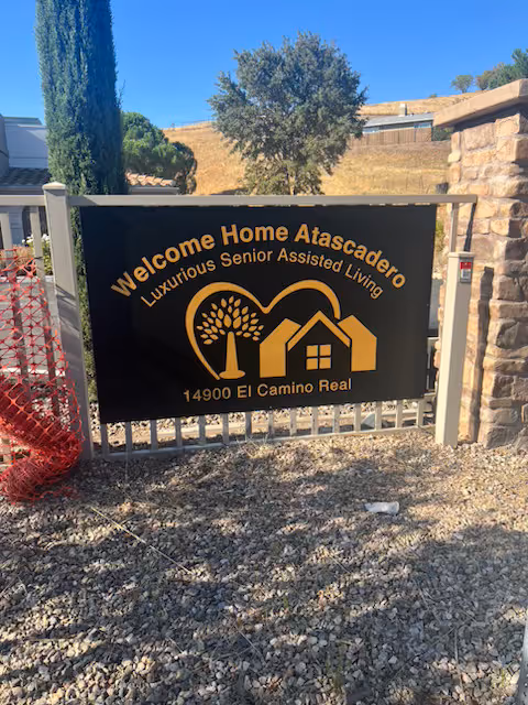 A black sign with gold lettering and graphics at the entrance of a senior living facility. The sign reads 'Welcome Home Atascadero Luxurious Senior Assisted Living 14900 El Camino Real' and features a graphic of a tree and two houses. The sign is mounted on a metal fence with stone pillars on either side, with a tree and dry hillside visible in the background under a clear blue sky.