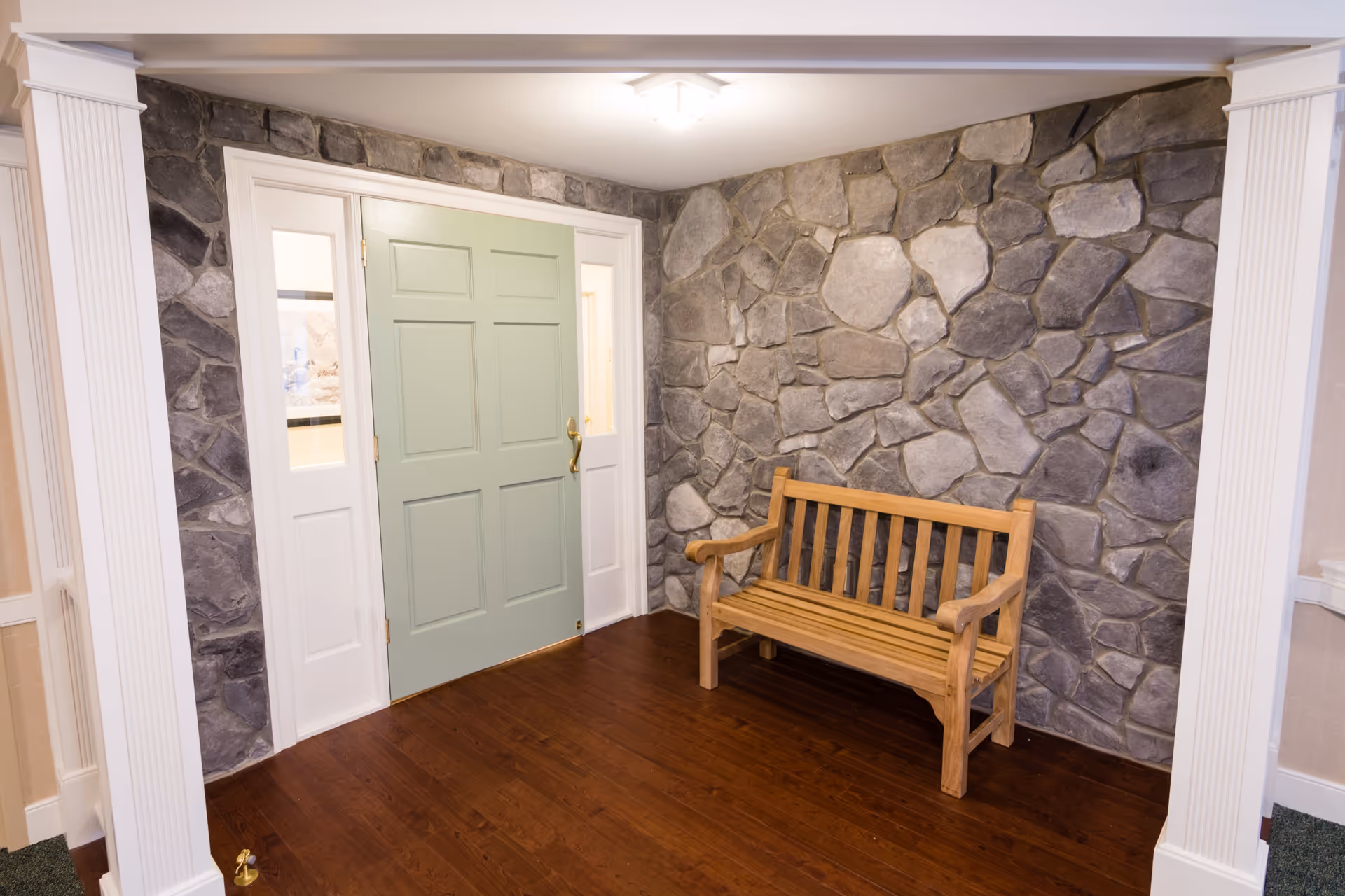 Interior view of a room with a stone accent wall, a light green door with glass panels on either side, a wooden bench, and hardwood flooring. The room has white trim and columns framing the entrance.