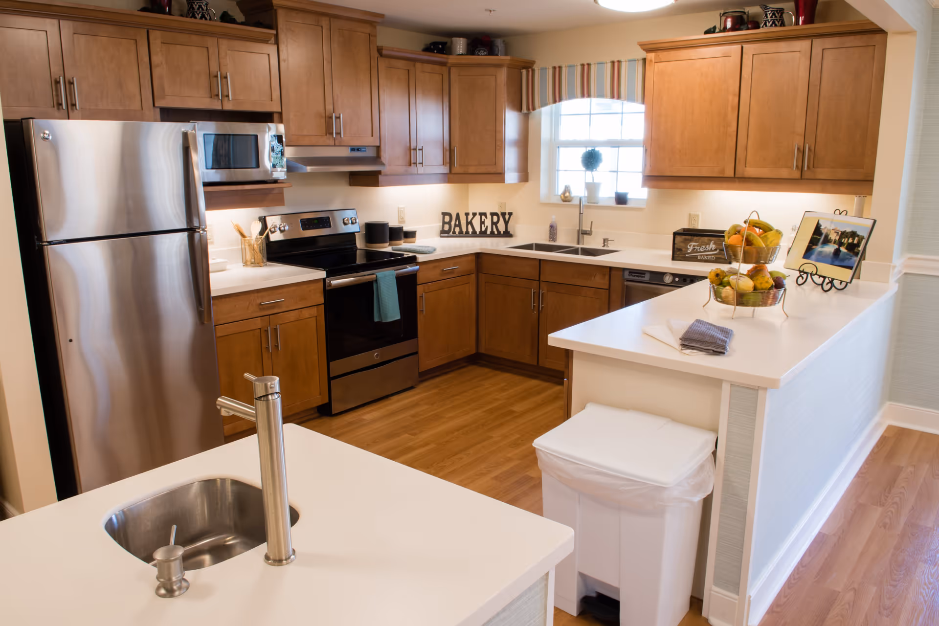 A modern kitchen with wooden cabinets, stainless steel refrigerator, stove, and microwave. The kitchen has white countertops, a double sink under a window with a small plant, and a kitchen island with a small sink and a trash bin underneath. There are decorative items including a fruit basket, a sign that says 'BAKERY', and a framed photo on the counter.