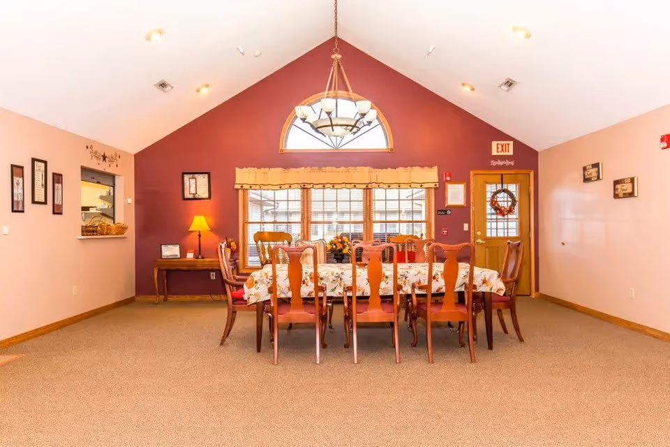 A dining room with a long table covered with a floral tablecloth and surrounded by wooden chairs. The room has a high vaulted ceiling with a large chandelier hanging above the table. There is a window with blinds and a valance on the far wall, a wooden door with a wreath, and a small table with a lamp and decorations on the left side. The walls are painted in light beige and dark red colors.
