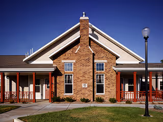 Front exterior view of a single-story brick and siding building with a peaked roof, red wooden porch railings, and a lamp post on the right side, under a clear blue sky.