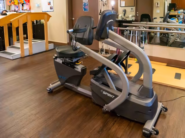 A recumbent exercise bike in a room with wooden flooring, a small set of stairs with handrails, a mirror on the wall, and various exercise equipment in the background.