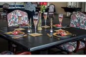 A dining table set for two with floral-patterned chairs. The table has gold-colored cutlery, glasses with pink liquid, napkins with decorative holders, and a vase with red roses in the center. The background shows more chairs and tables in a softly lit room.