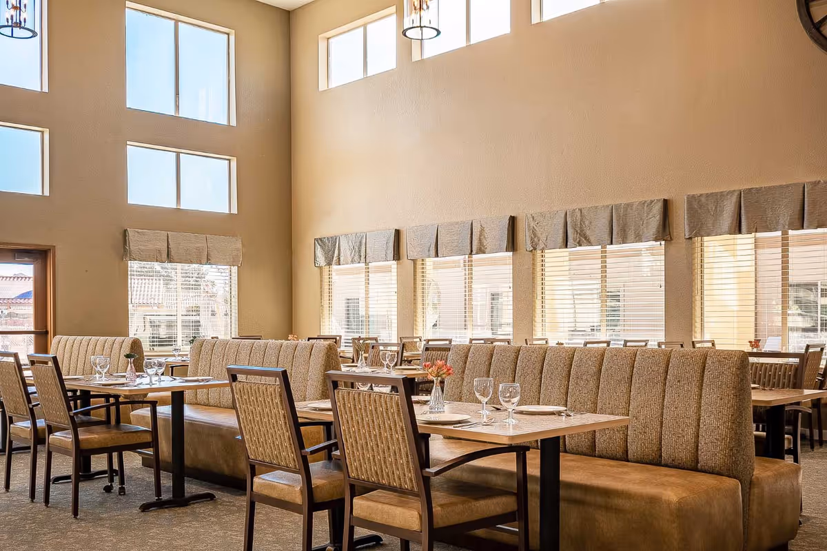 A bright dining room with large windows and beige walls. The room features long cushioned bench seating along the windows and wooden chairs around tables set with plates, glasses, and small flower vases.