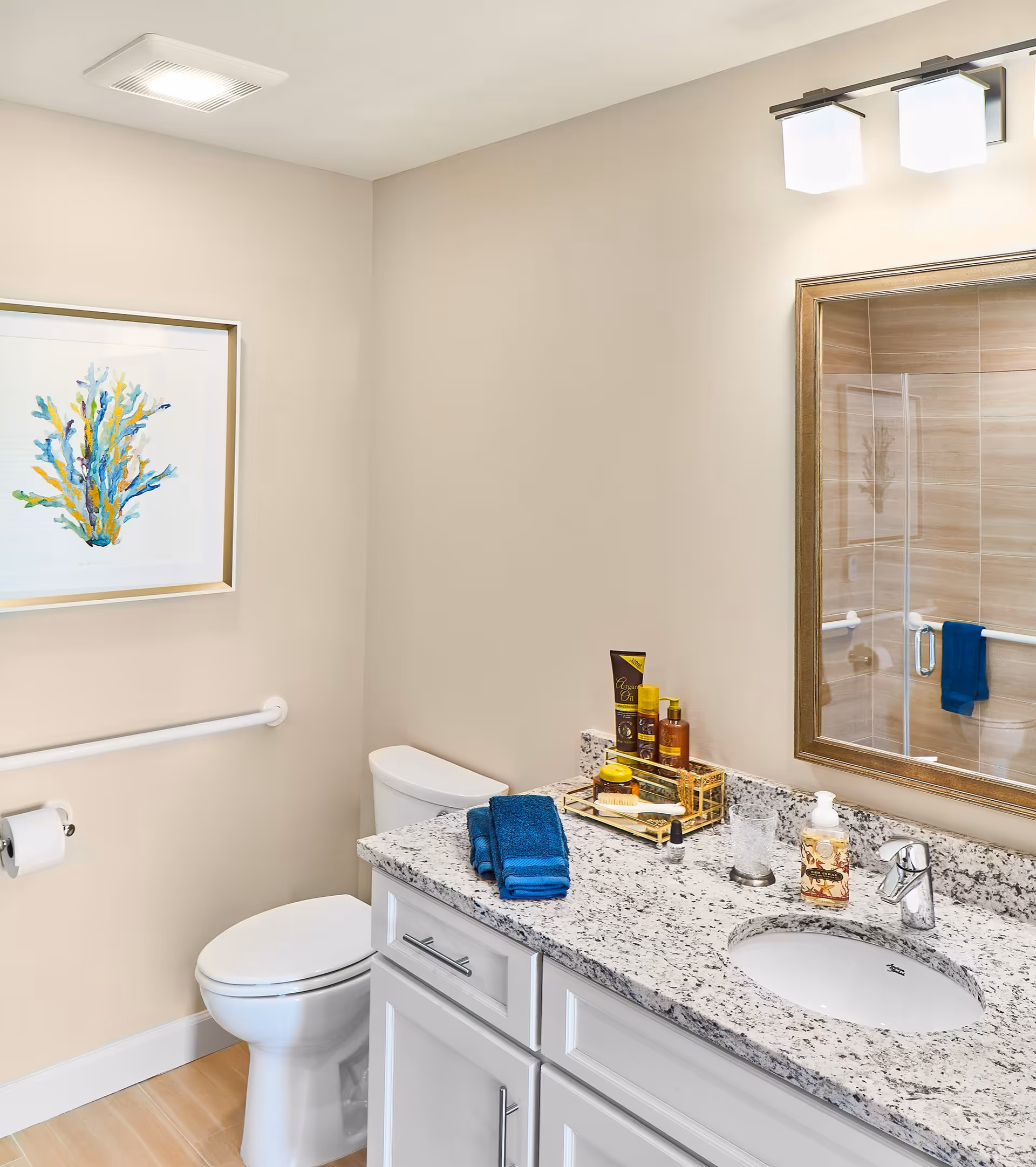 A clean and modern bathroom featuring a white toilet, a granite countertop with a built-in sink, and a large mirror with a wooden frame. On the countertop are neatly folded blue towels, various toiletries, and a soap dispenser. The walls are painted beige, and there is a framed artwork of colorful coral on the wall. A glass-enclosed shower with beige tiles and a blue towel hanging inside is reflected in the mirror.