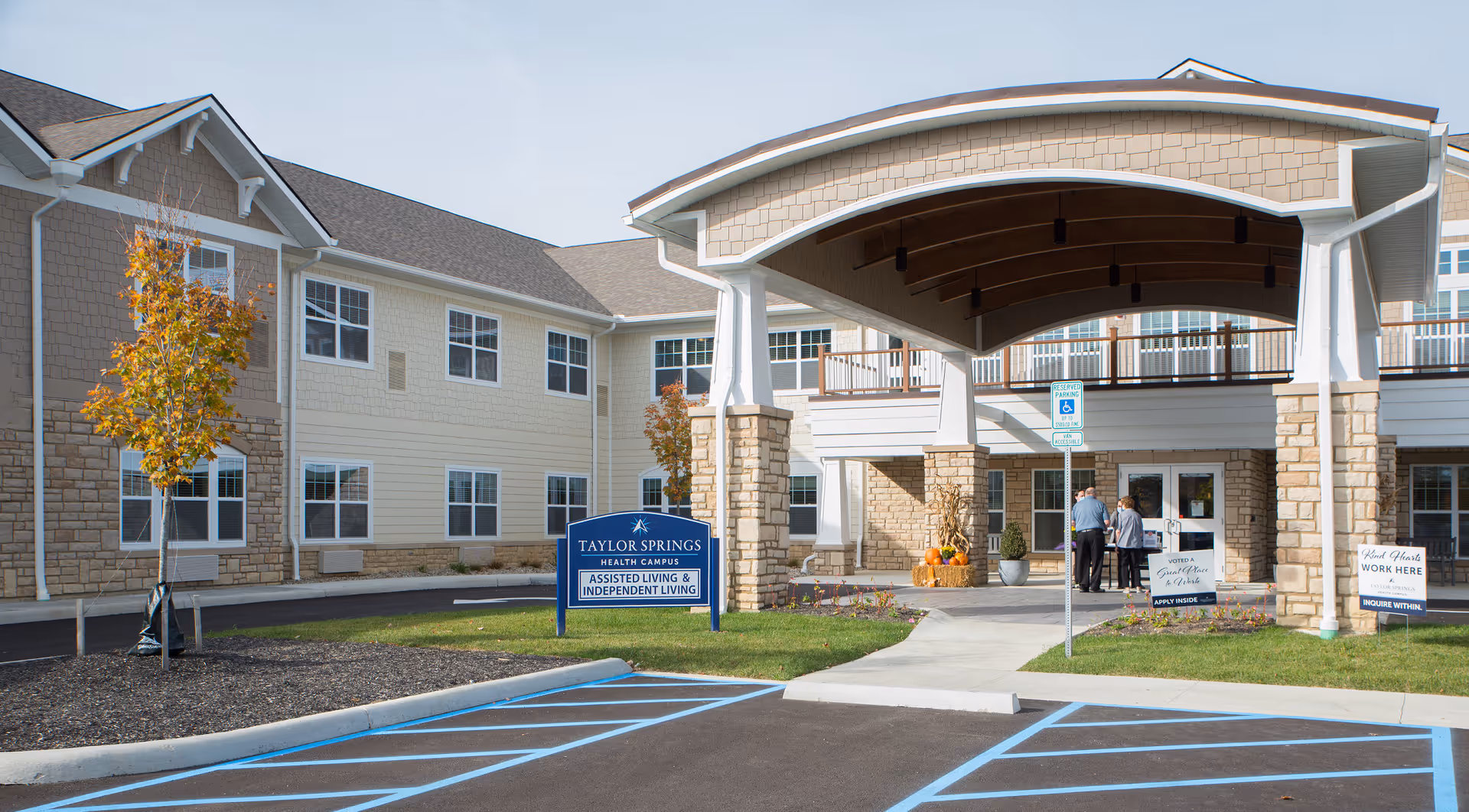 Exterior view of Taylor Springs Health Campus building entrance with a covered drop-off area, two people standing near the entrance, a sign indicating assisted living and independent living, and a parking lot with handicap spaces in the foreground.
