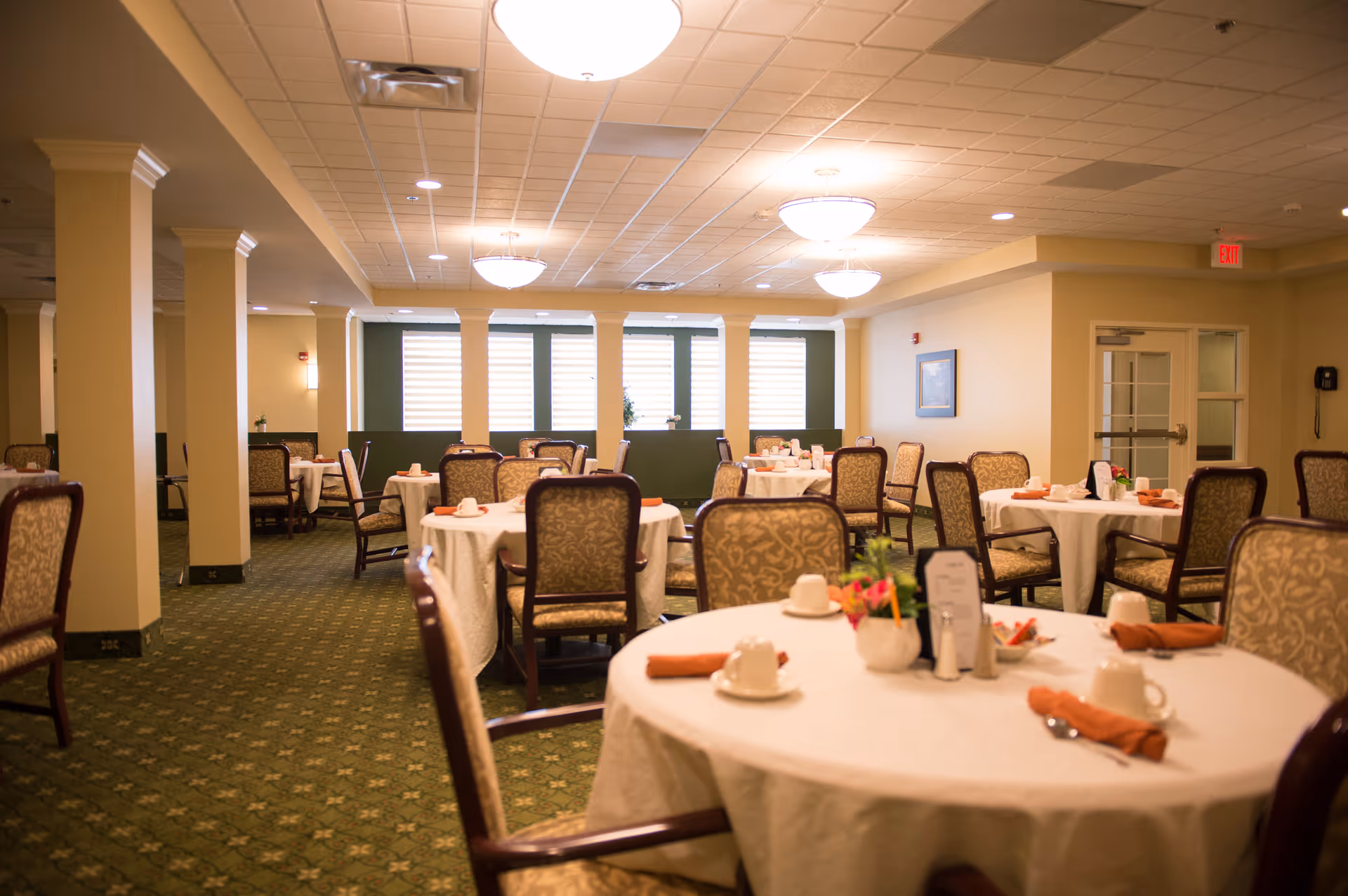 A spacious dining room with round tables covered in white tablecloths, each set with cups, saucers, and orange napkins. The room has patterned carpet flooring, beige walls, multiple upholstered chairs, and large windows with blinds letting in natural light. Ceiling lights provide additional illumination.