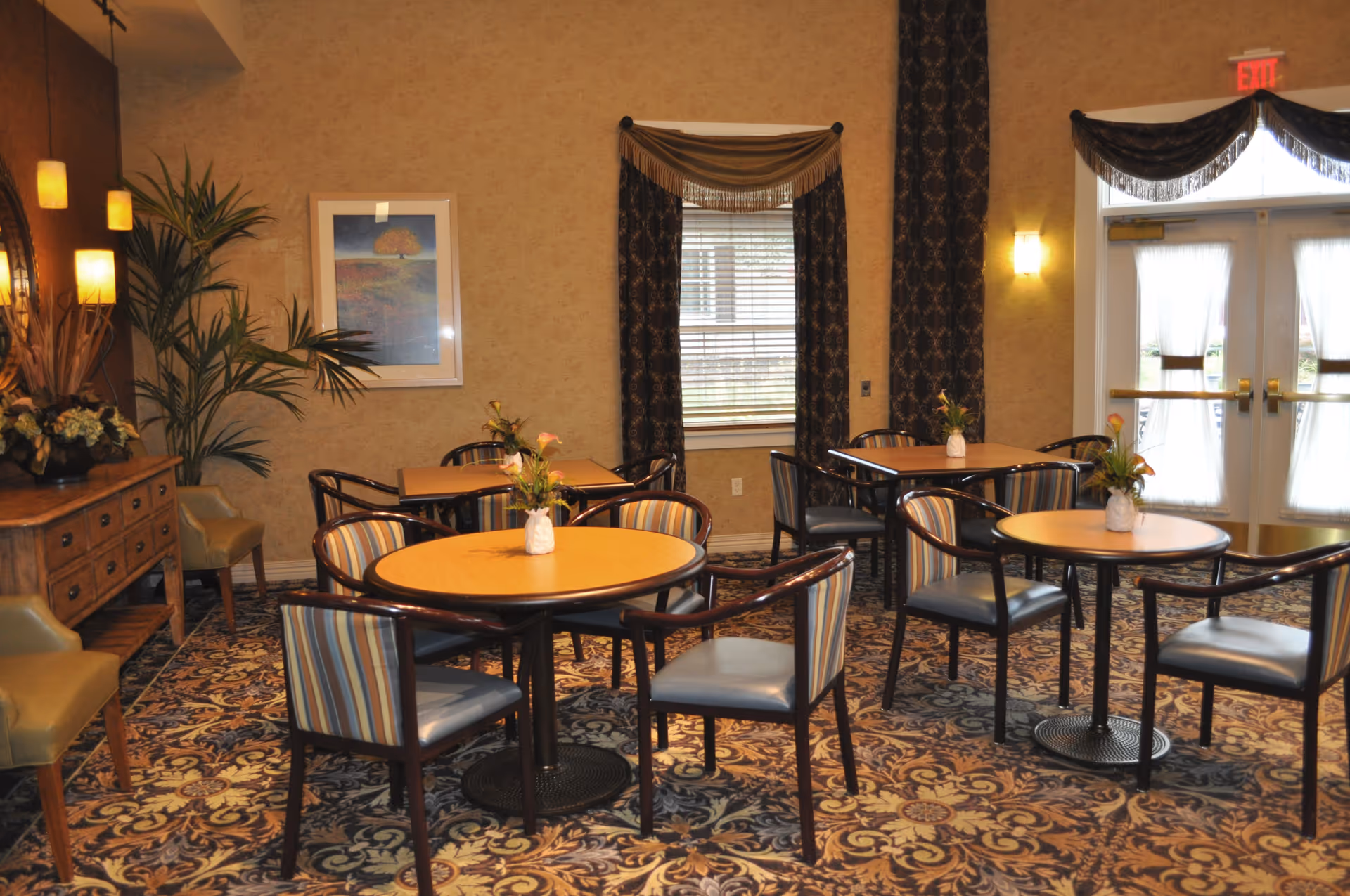 A cozy dining area with several round and square wooden tables surrounded by chairs with striped cushions. The room features patterned carpet, beige walls, a window with dark curtains, a sideboard with decorative plants and lamps, and double doors with sheer curtains letting in natural light.