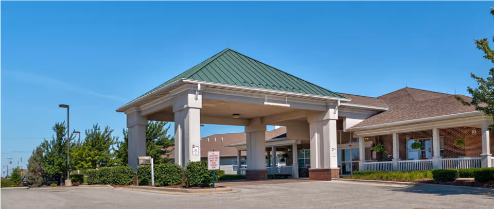 Exterior view of Liberty Village of Clinton showing the entrance with a covered drop-off area supported by large columns, a green metal roof, brick walls, and surrounding greenery under a clear blue sky.