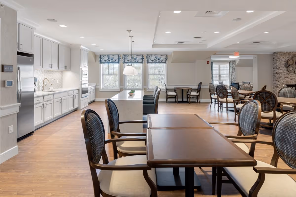 A bright and spacious dining area in a senior living facility featuring multiple wooden tables with cushioned chairs. The room has large windows with patterned valances allowing natural light to fill the space. On the left side, there is a modern kitchen area with white cabinetry, stainless steel appliances, and a large island with pendant lighting above. The floor is wood, and the walls are painted in light neutral tones.
