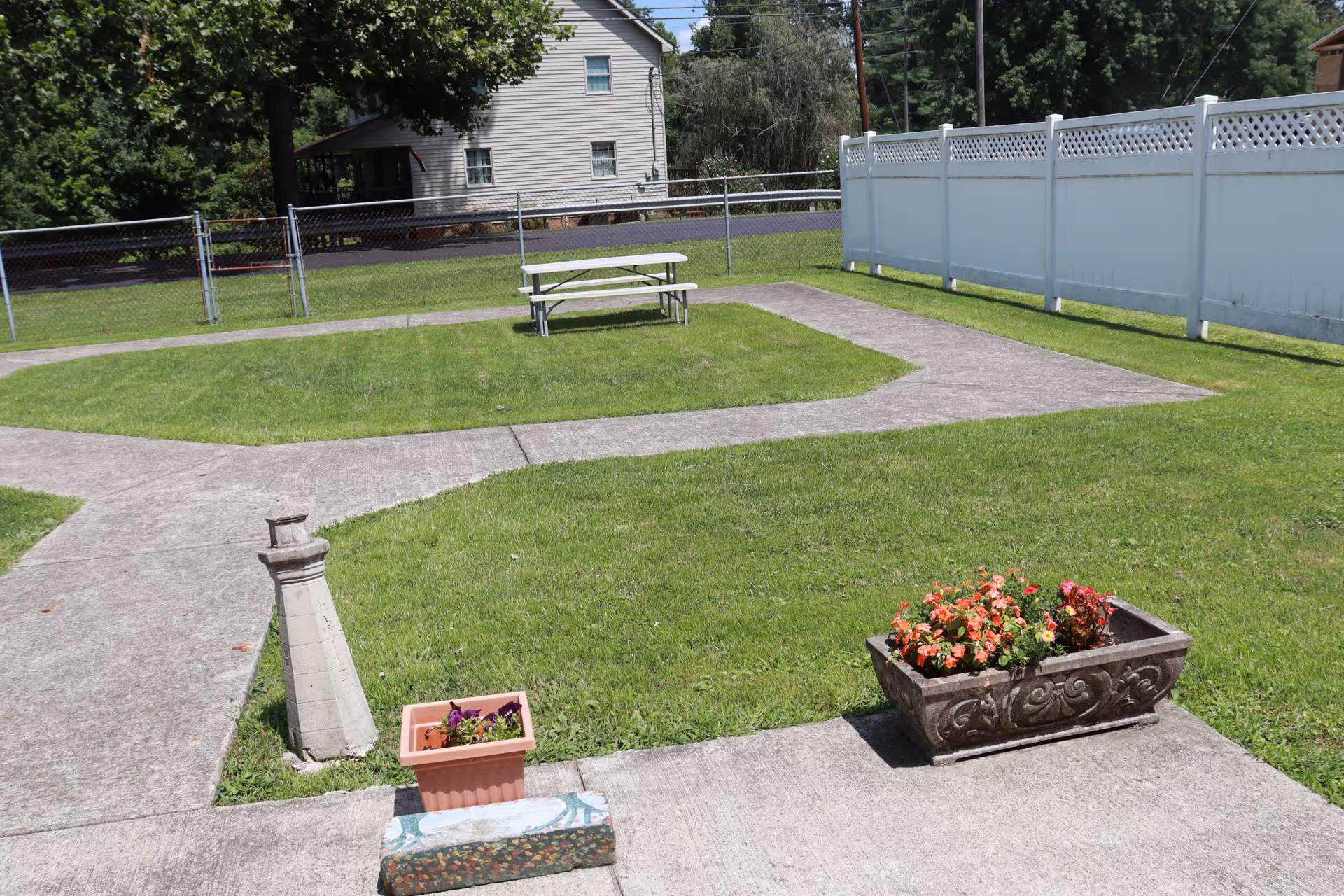 Outdoor area with green grass, concrete walking paths, a white picnic table, two flower pots with colorful flowers, a small lighthouse decoration, and a white fence on the right side. A house and trees are visible in the background.