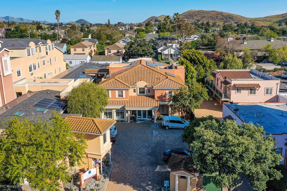 Aerial view of Avila Senior Living At Downtown SLO showing a large building with terracotta roof tiles surrounded by trees and other residential buildings, with hills in the background under a clear blue sky.