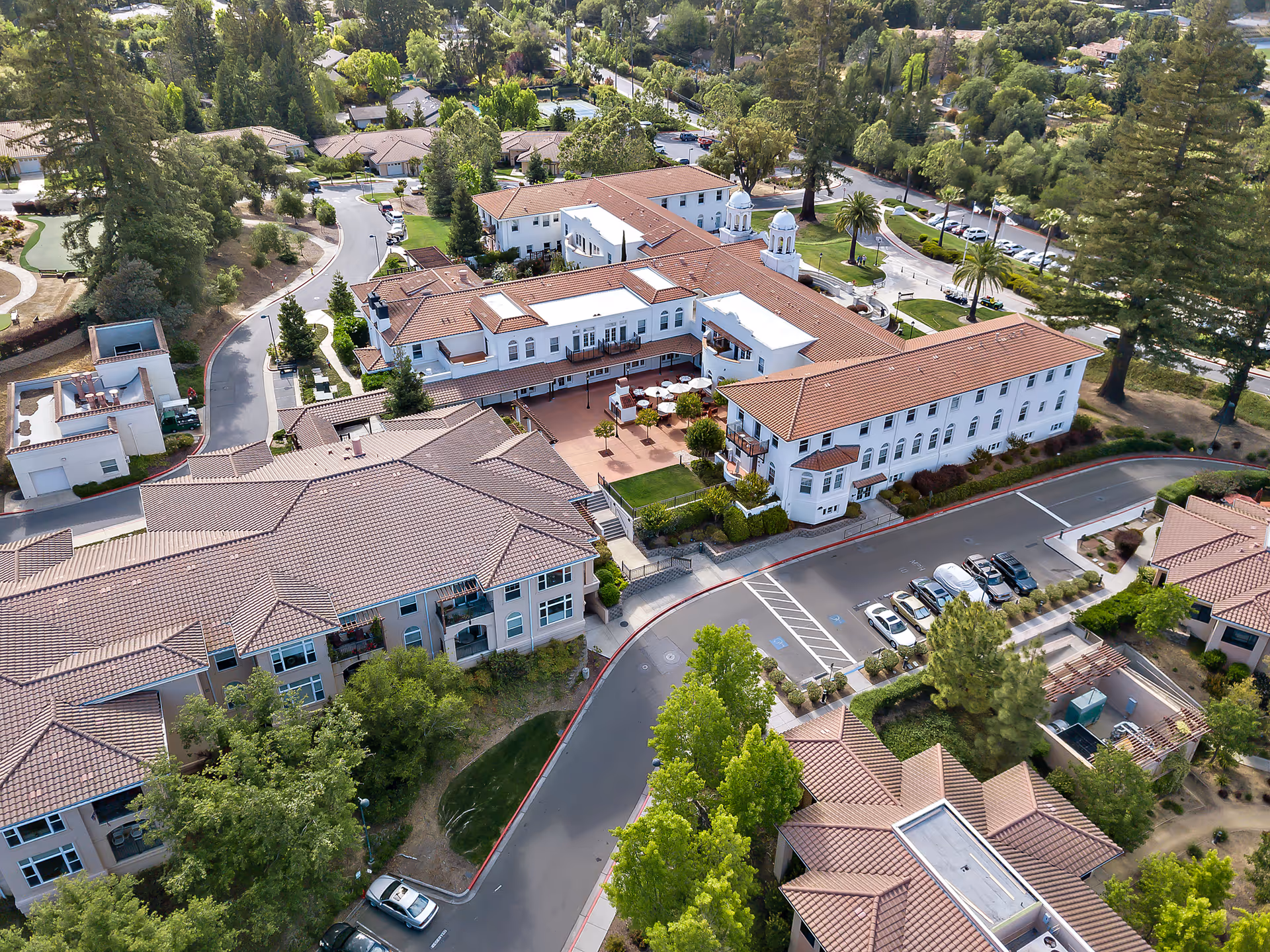 Aerial view of Saratoga Retirement Community Assisted Living showing multiple connected buildings with red-tiled roofs, surrounded by trees, parking areas, and landscaped grounds.