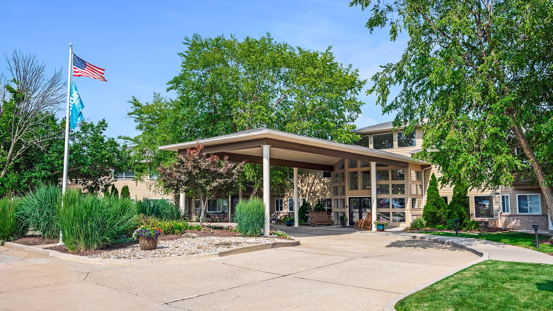 Exterior view of Holiday Palmer Hills facility entrance with a covered drop-off area, surrounded by green trees and landscaping. Two flagpoles display the American flag and another blue flag. The building has large windows and a beige facade.
