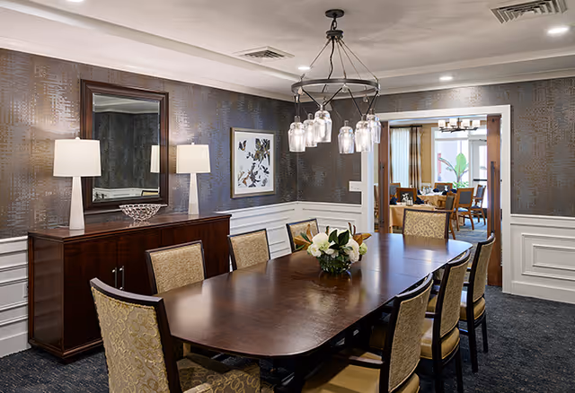 A formal dining room with a long wooden table surrounded by eight upholstered chairs. The room features dark textured wallpaper with white wainscoting, a wooden sideboard with two table lamps and a decorative bowl, a large mirror, and framed artwork on the walls. A modern chandelier with multiple glass shades hangs above the table. Through an open doorway, another room with additional seating and a chandelier is visible.