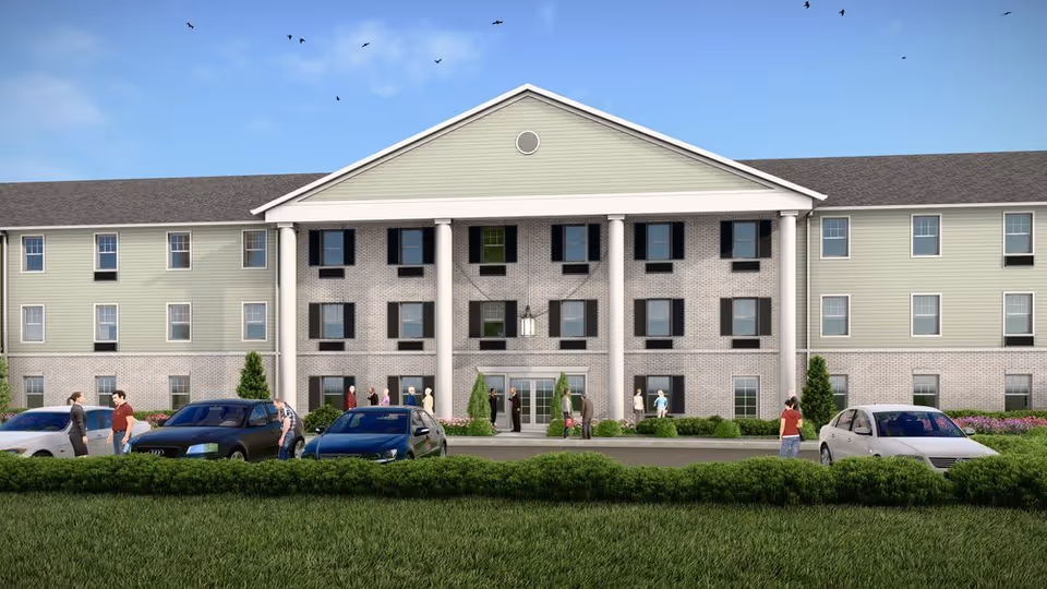 Front exterior view of a three-story senior living facility building with a central entrance flanked by white columns. Several people are standing and walking near the entrance and parked cars are visible in front. The building has a combination of light green siding and brickwork with multiple windows and a clear blue sky with birds flying above.