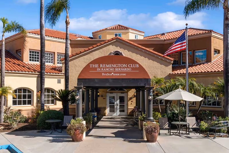 Front exterior view of The Remington Club senior living facility in Rancho Bernardo, featuring a beige building with red tile roofing, palm trees, an American flag, and outdoor seating with tables and umbrellas.