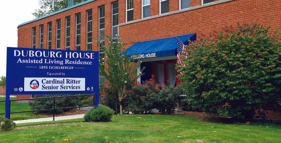 Exterior view of Dubourg House Assisted Living Residence, a red brick building with a blue awning over the entrance. There is a large blue sign in front that reads 'Dubourg House Assisted Living Residence, 5890 Eichelberger, Operated by Cardinal Ritter Senior Services.' The area is landscaped with green grass, bushes, and small trees.