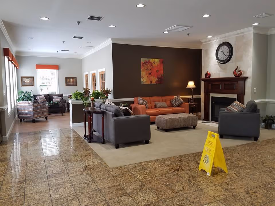 A spacious and well-lit living room area with a tiled floor and a carpeted seating section. The seating area includes a brown sofa, two gray armchairs, and an ottoman, arranged around a fireplace with a clock above it. There is a side table with a lamp and decorative items on the mantel. In the background, there is another seating area with patterned armchairs and a small table near windows with orange valances. Several plants and framed pictures decorate the space. A yellow caution sign is placed on the tiled floor.