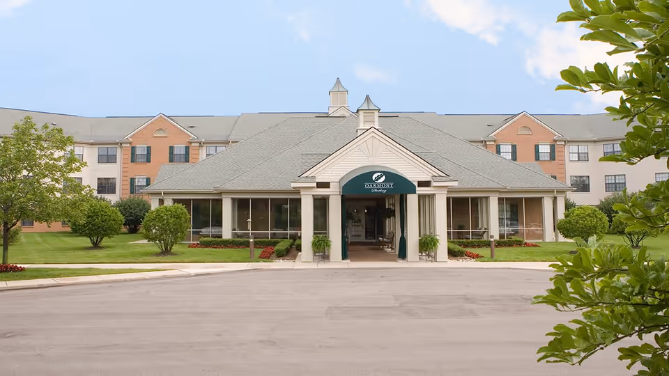 Front exterior view of Oakmont Sterling Senior Community building with a covered entrance, manicured lawn, trees, and clear sky.