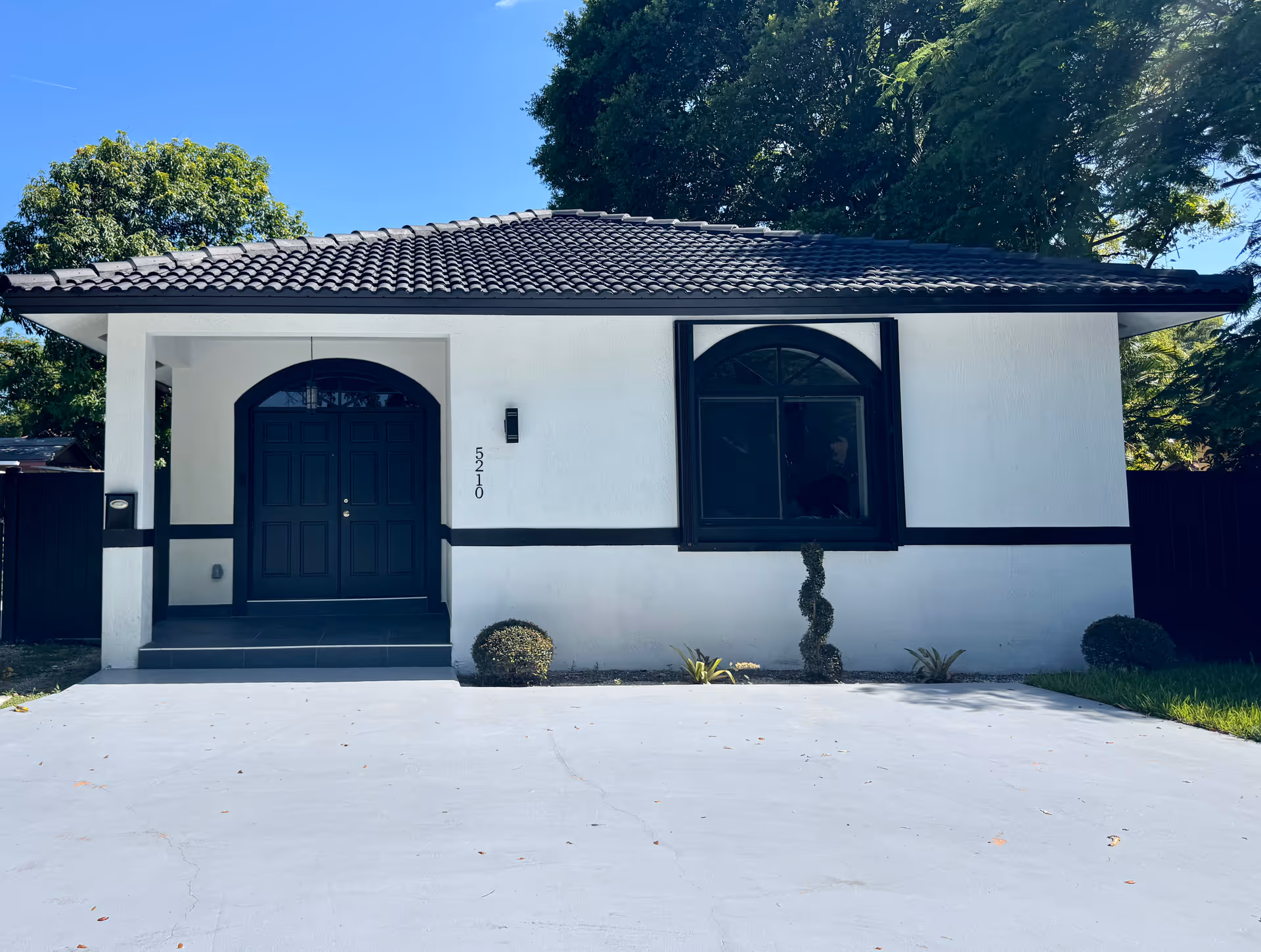 Front exterior view of a single-story building with white walls and black trim. The building has a tiled roof, a black double door entrance with an arched top, and a large window with black framing. There are small bushes and plants in front of the building, and trees in the background under a clear blue sky.