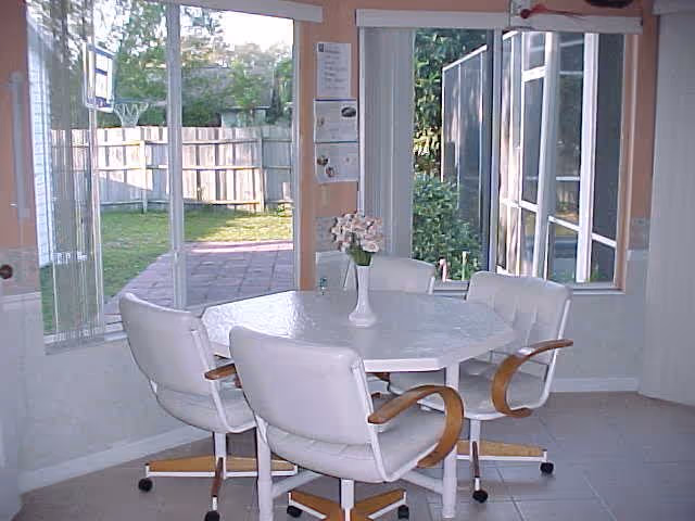 A small dining area with a round white table and four white cushioned chairs with wooden armrests on wheels. A white vase with flowers is placed on the table. Large windows and a sliding glass door provide a view of a fenced backyard with grass and a paved patio area.