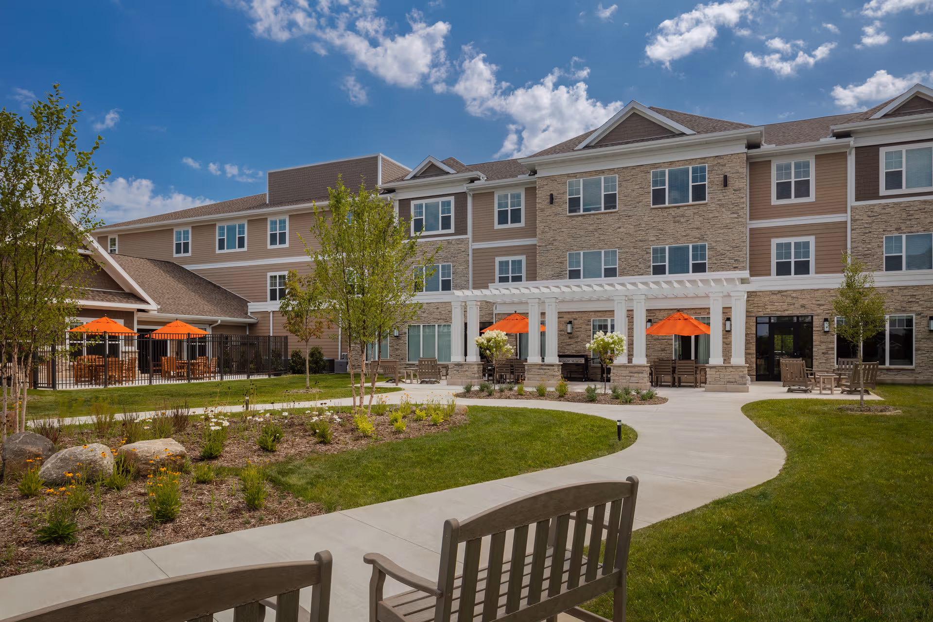 Outdoor courtyard area of a senior living facility with a paved walkway, green lawn, small trees, and landscaped flower beds. The building has three stories with beige and stone exterior walls. There are several seating areas with wooden benches and tables shaded by orange umbrellas under a white pergola and near a fenced patio area.