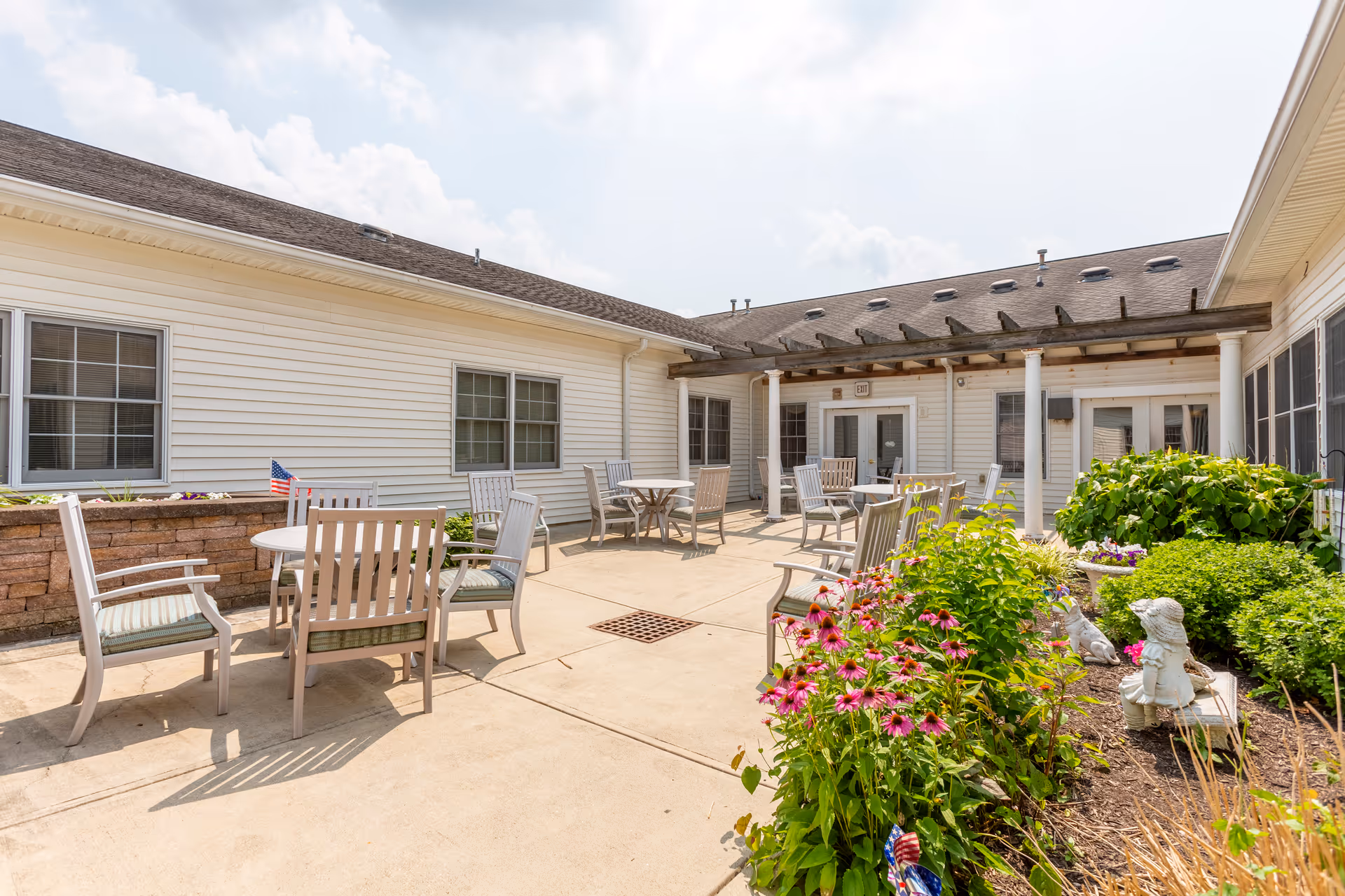 Outdoor courtyard area at Independence Village of Avon (Prestwick) featuring several round tables with chairs arranged on a concrete patio. The space is surrounded by white buildings with windows and a pergola overhead. There are flower beds with green shrubs and blooming pink flowers along one side of the patio.