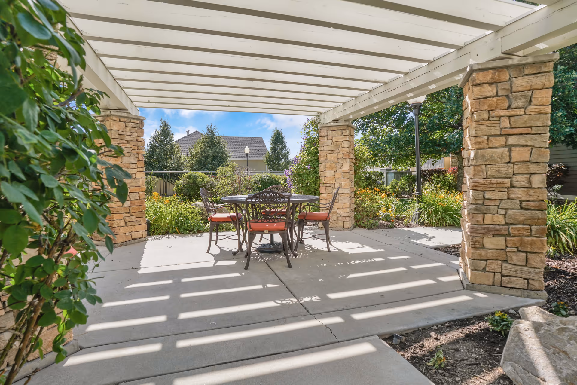 Outdoor patio area with a white pergola supported by stone pillars, featuring a round metal table and four chairs with red cushions, surrounded by greenery and flowers under a blue sky.
