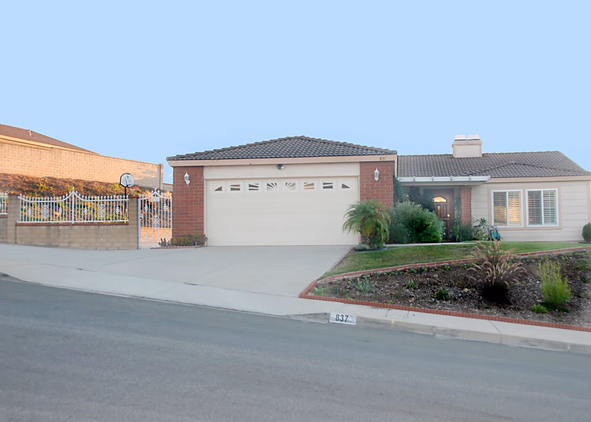 Front exterior view of a single-story residential building with a tiled roof, a two-car garage with a white door, a small garden with plants and shrubs, and a gated side yard.