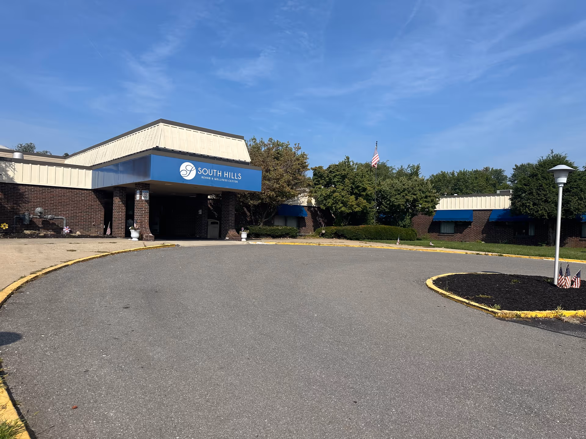 Exterior view of South Hills Rehab & Wellness Center building with a circular driveway, blue awnings, and an American flag on a flagpole surrounded by trees under a clear blue sky.