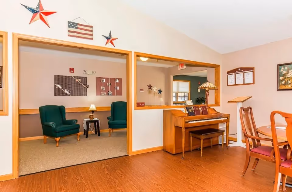 Interior view of a senior living facility room with a wooden piano, a piano bench, and a small table lamp on top. To the right, there is a round wooden table with chairs. Through a large window opening in the wall, two green upholstered armchairs and a small side table with a lamp are visible in an adjacent room. The walls are decorated with patriotic star decorations and framed artwork.