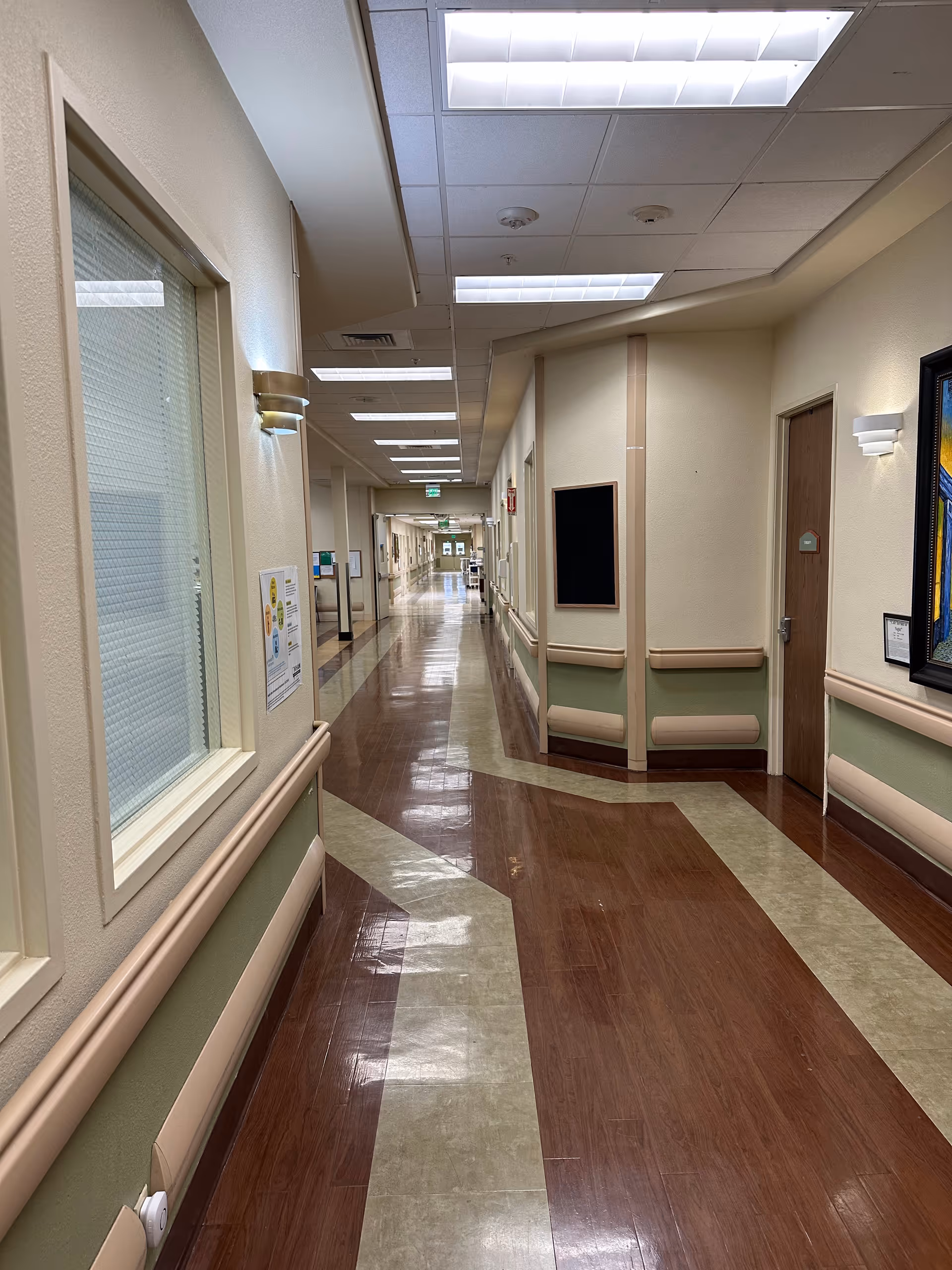 A long, clean hallway inside a nursing and rehabilitation center with polished brown and beige patterned flooring, beige walls with green accents, handrails along the walls, overhead fluorescent lighting, and several closed doors and windows along the corridor.