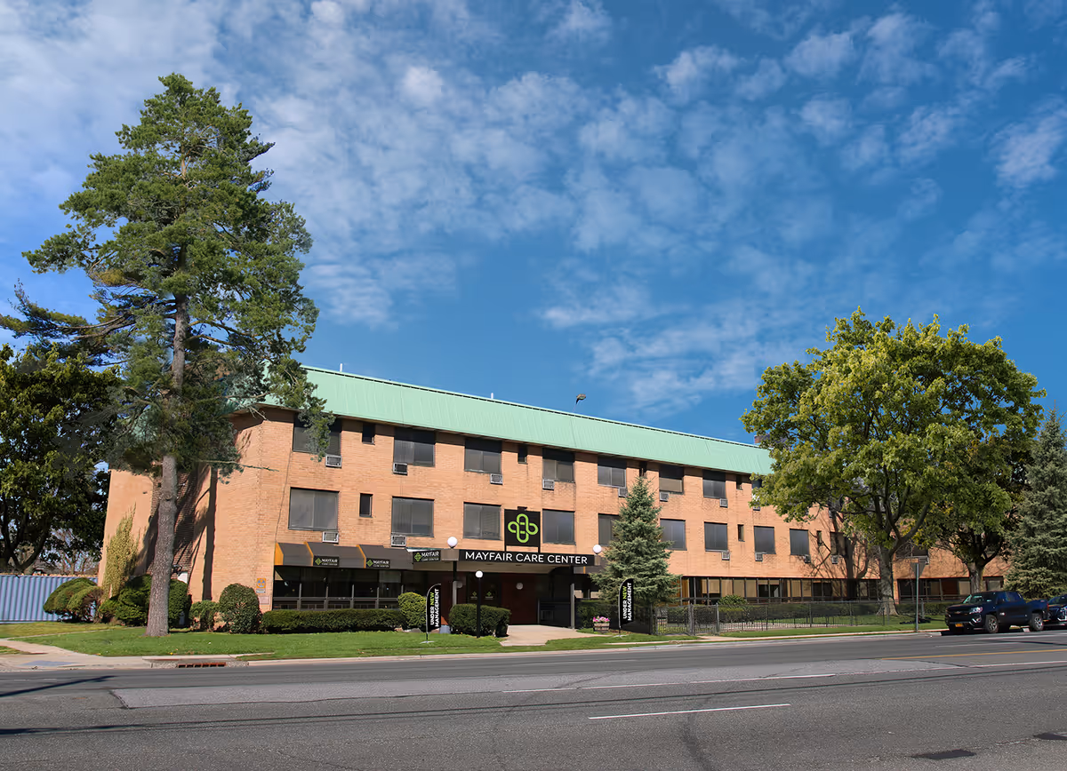 Exterior view of Mayfair Care Center, a three-story brick building with a green roof, surrounded by trees and bushes under a partly cloudy blue sky. There are a few parked cars along the street in front of the building.