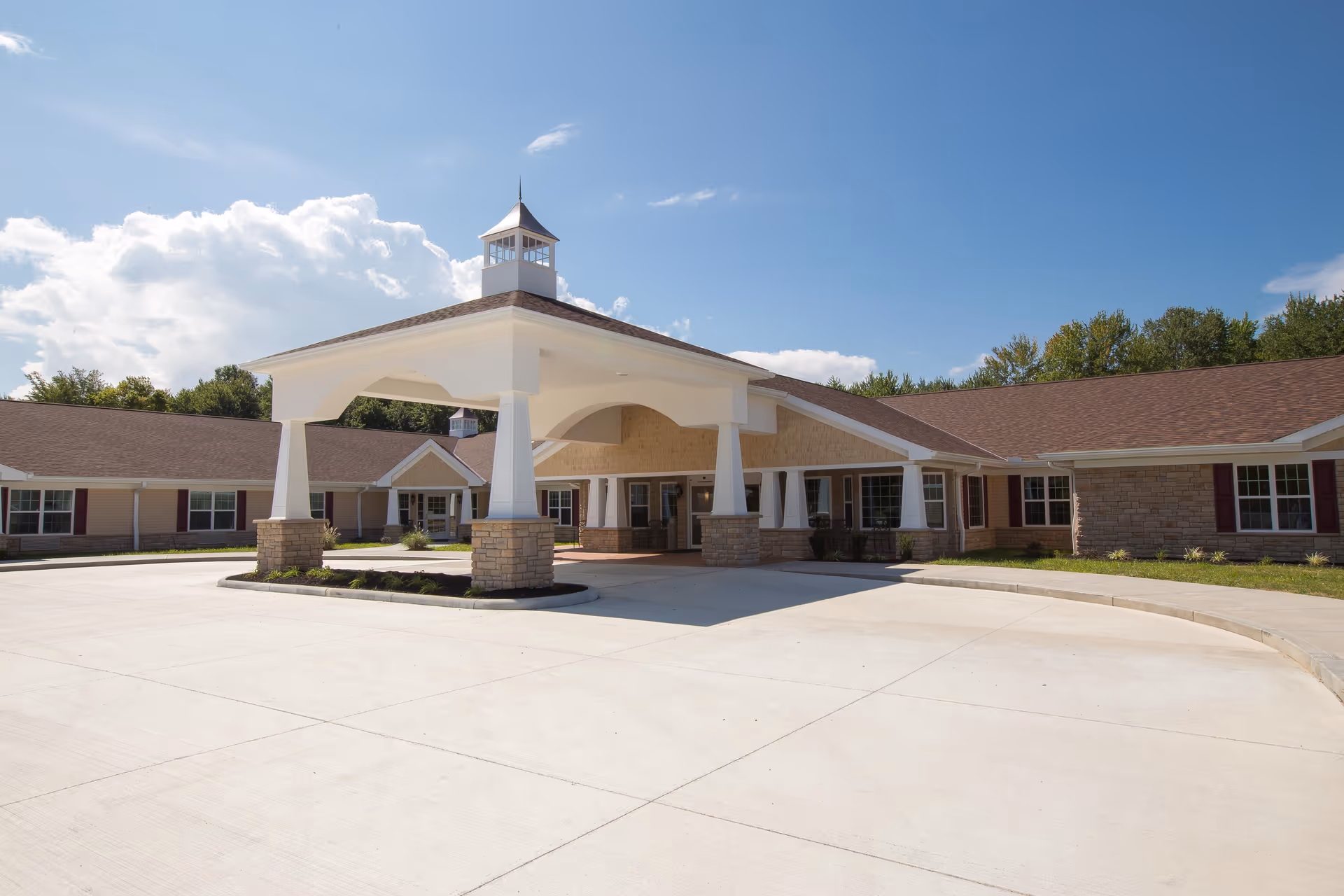 Exterior view of Siena Gardens Rehabilitation & Transitional Care facility showing a large covered entrance with white pillars and a cupola on the roof, surrounded by a circular driveway and a clear blue sky.