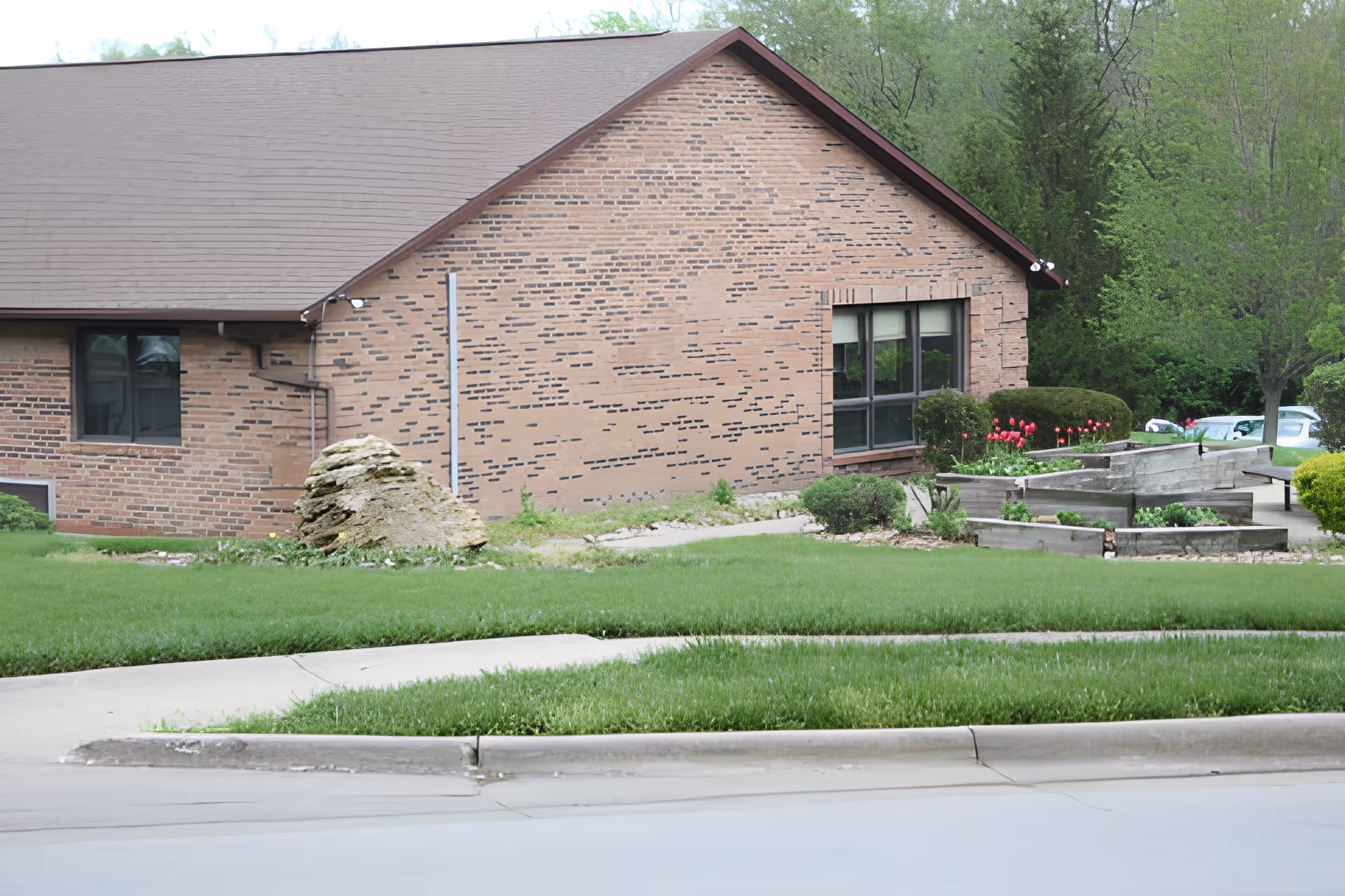 Exterior side view of a brick building with a sloped roof, surrounded by green grass, bushes, and trees. There is a large rock near the building and a garden area with wooden raised beds and red flowers. A sidewalk and street are visible in the foreground.
