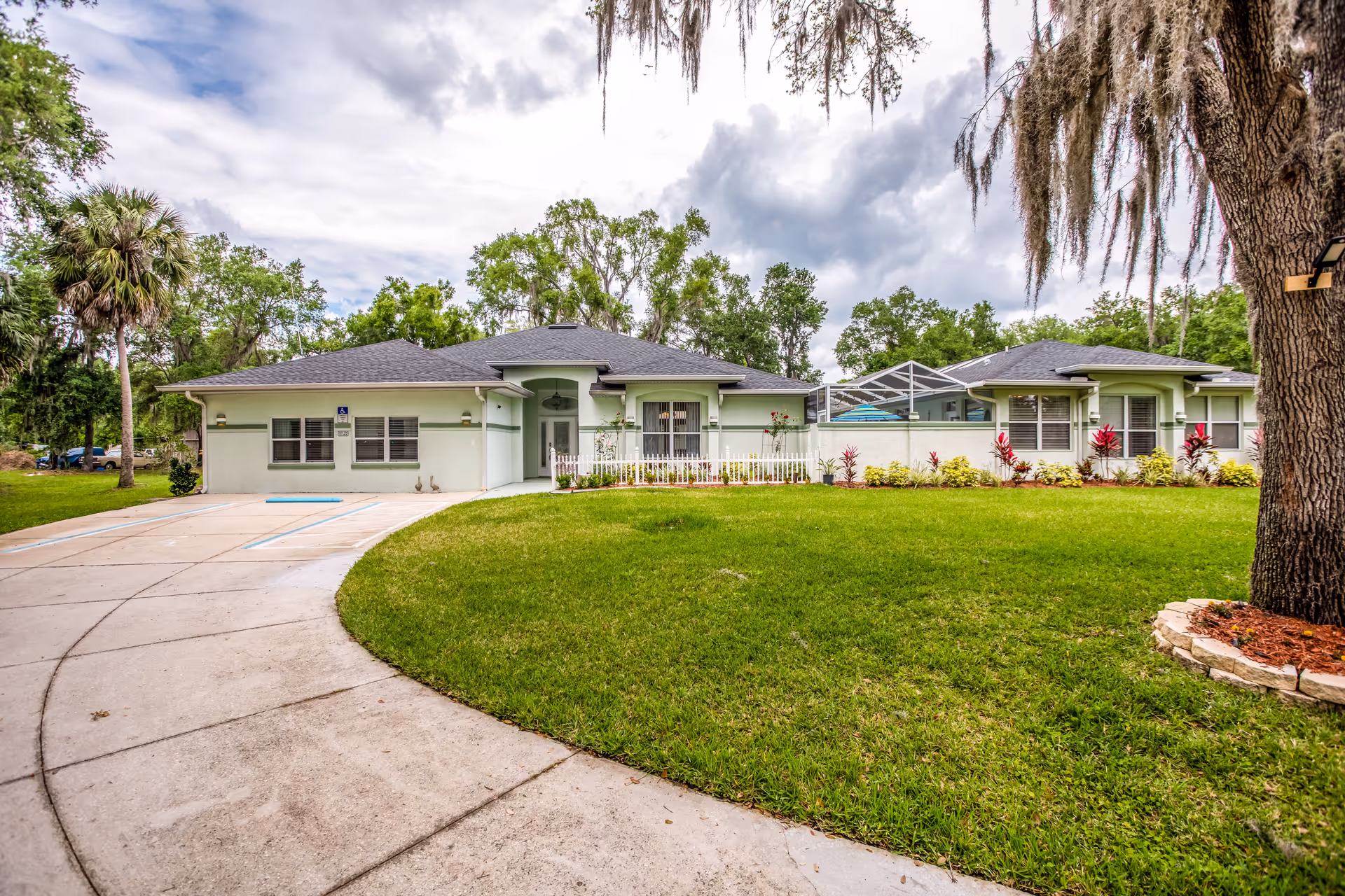 Front exterior view of a single-story assisted living home with a curved driveway, green lawn, and trees surrounding the property under a cloudy sky.