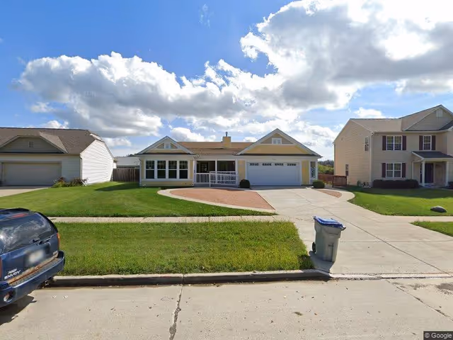 A single-story suburban house with a front porch and attached two-car garage, surrounded by a well-maintained lawn. The sky is partly cloudy with bright sunlight. A trash bin is placed at the edge of the driveway, and a black vehicle is parked on the street in front of the house.
