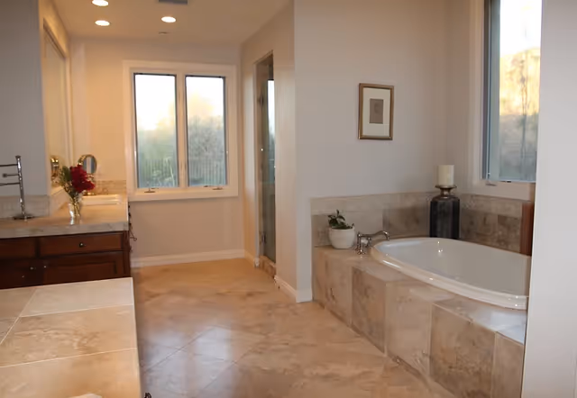 A spacious bathroom with beige tiled floors and walls. There is a built-in bathtub with a faucet, a potted plant, and a candle holder next to it. A large window is above the bathtub letting in natural light. On the left side, there is a countertop with a sink, a mirror, and a vase with red flowers. In the background, there is a glass-enclosed shower and another window.