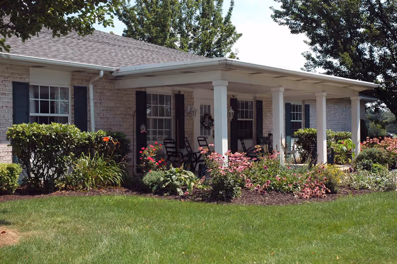 Exterior view of a single-story brick building with a covered porch featuring several rocking chairs. The porch is supported by white columns and surrounded by well-maintained flower beds and green shrubs. The lawn in front is neatly mowed and there are trees in the background.