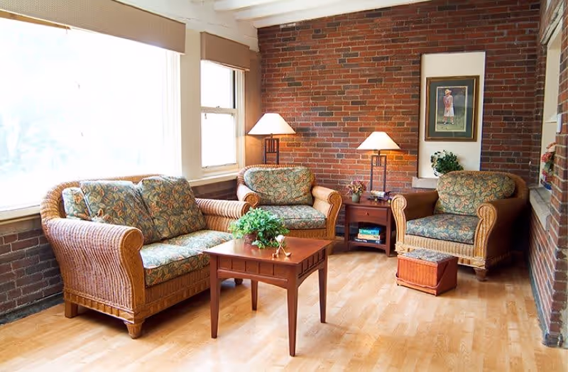 A cozy living room with wicker furniture including a sofa and two armchairs with floral cushions. There is a wooden coffee table with a green plant on it, two side tables with lamps, a framed picture on the brick wall, and a small ottoman on a light wooden floor. Large windows let in natural light.