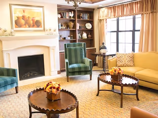 A cozy living room with a white fireplace, a painting above it, built-in wooden bookshelves filled with decorative items, two green armchairs, a yellow sofa, a window with beige curtains, and two round wooden coffee tables with flower arrangements.