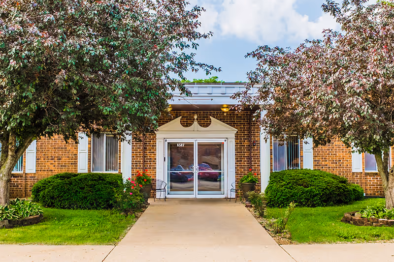 Front entrance of a brick building with white framed double glass doors, flanked by two chairs and large potted plants. There are two trees and well-maintained bushes on either side of the concrete walkway leading to the entrance under a partly cloudy sky.