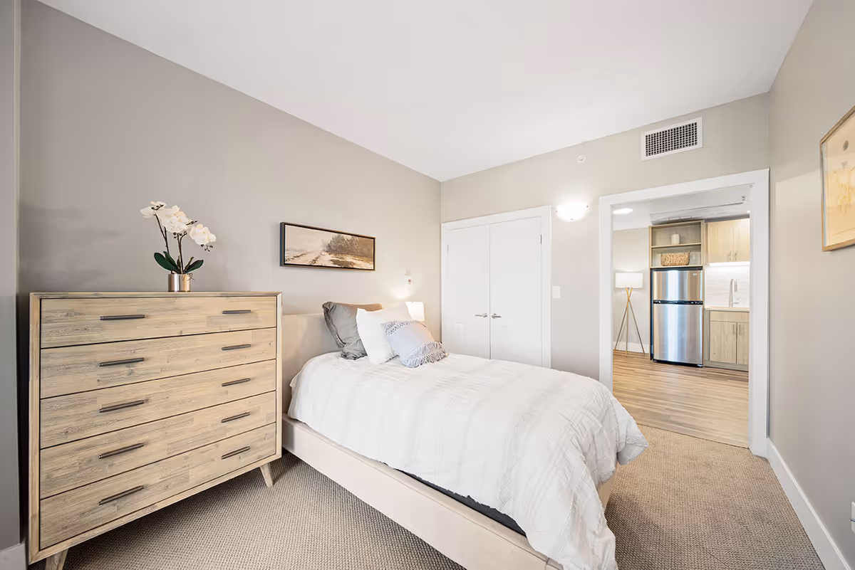 A neatly arranged bedroom with a bed covered in white bedding and multiple pillows. To the left of the bed is a wooden dresser with a potted orchid on top. A framed landscape picture hangs on the wall above the bed. The room has beige walls and carpeted flooring. Through an open doorway, a kitchen area with wooden cabinets and a stainless steel refrigerator is visible.