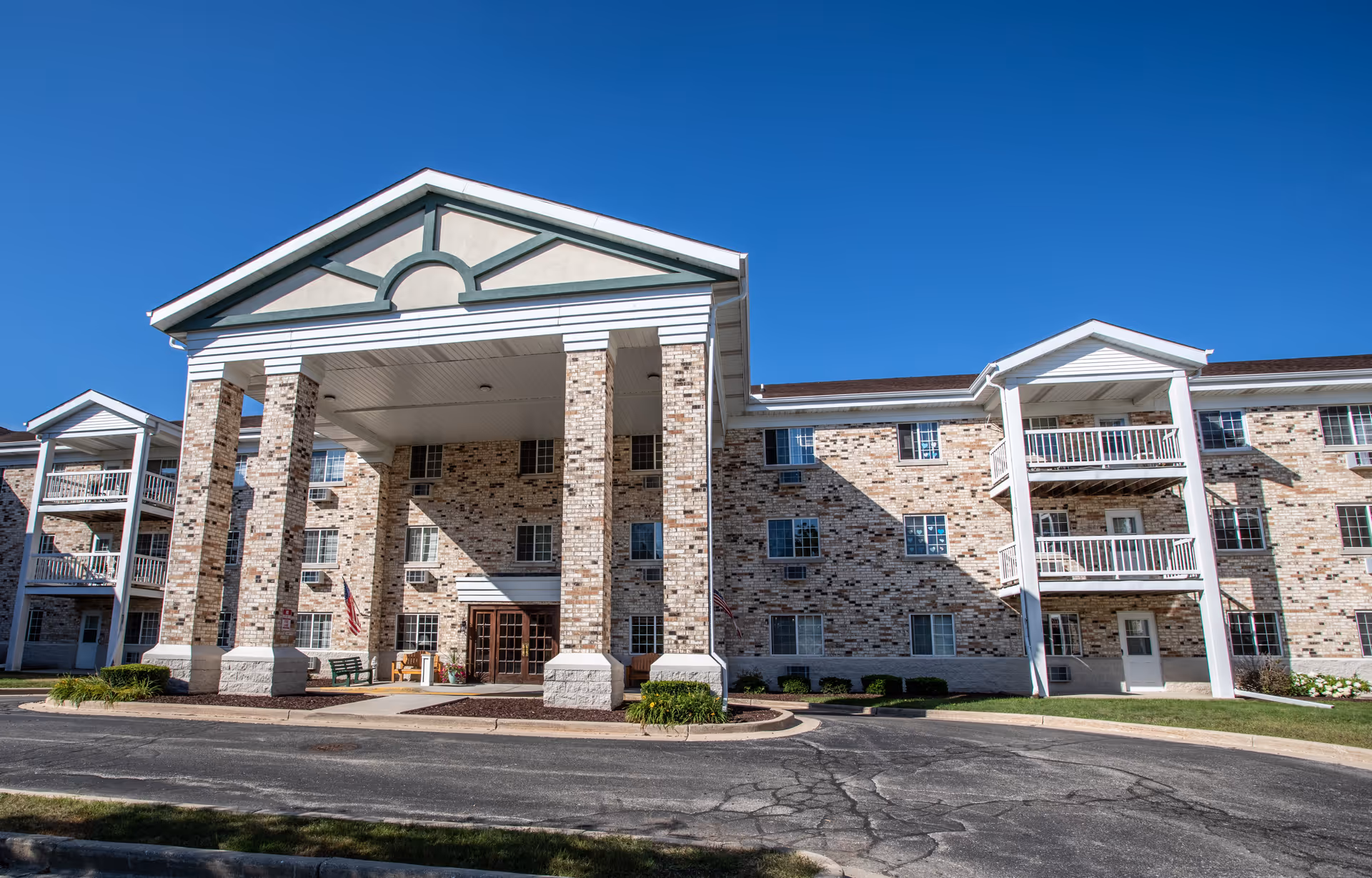 Front exterior view of a three-story senior living facility building with a large covered entrance supported by brick columns, balconies on the sides, and a clear blue sky above.