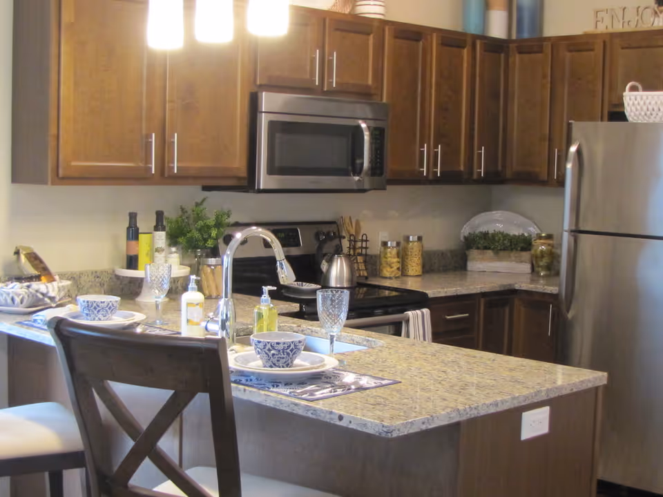 Modern kitchen with granite countertops, wooden cabinets, stainless steel microwave and refrigerator. The kitchen island has two place settings with blue and white bowls, plates, glasses, and silverware. There are various kitchen items and decorative plants on the countertops.