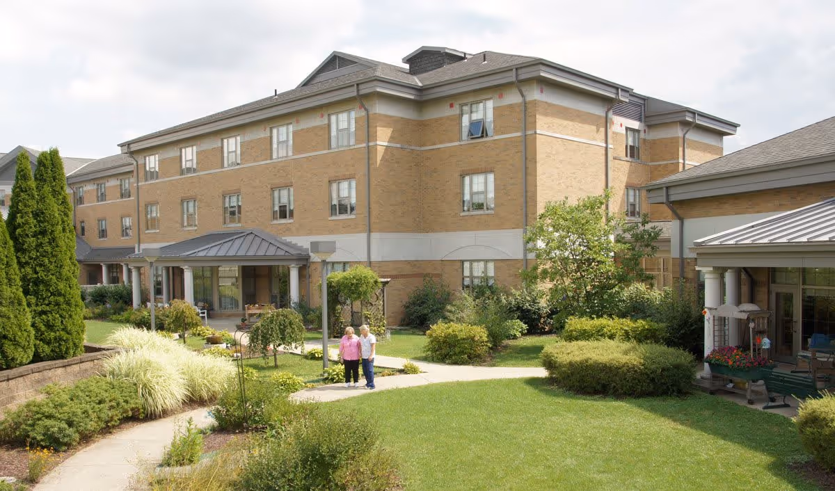Exterior view of a three-story brick building with multiple windows, surrounded by well-maintained green lawns, bushes, and trees. Two elderly people are standing on a paved walkway in the garden area. The building has a covered entrance with columns and a metal roof.