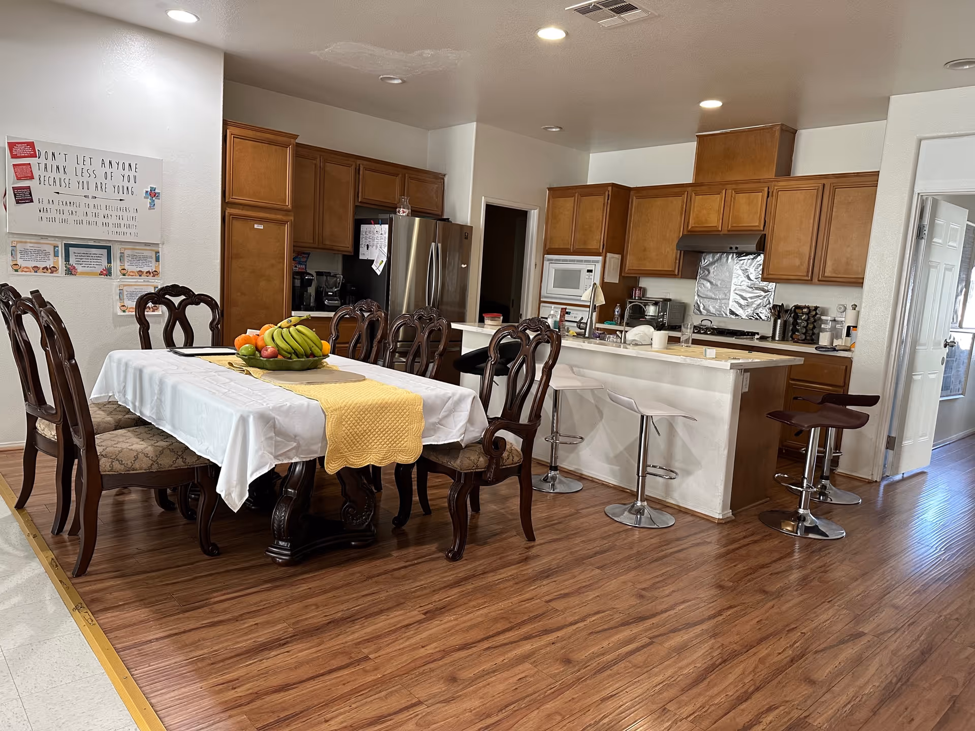 A spacious kitchen and dining area with wooden cabinets, a stainless steel refrigerator, a microwave, and a kitchen island with bar stools. The dining table is covered with a white tablecloth and a yellow runner, and has a fruit bowl centerpiece. The floor is wooden, and there are motivational posters on the wall.