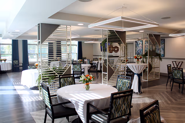 A bright and spacious dining room with round tables covered in white tablecloths, each adorned with a small floral centerpiece. The room features modern decor with geometric light fixtures hanging from the ceiling and decorative partition panels. Chairs with patterned upholstery surround the tables, and large windows allow natural light to fill the space.