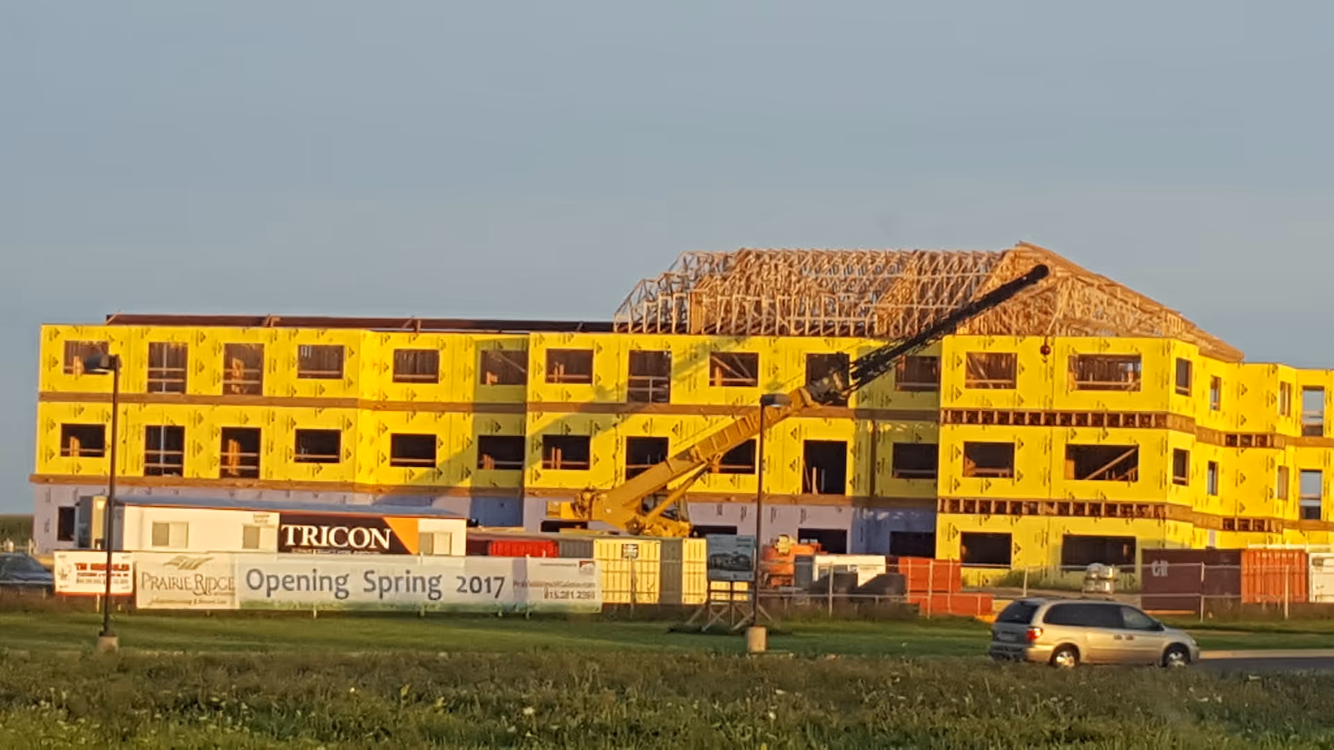 A large multi-story building under construction with yellow sheathing on the exterior walls and wooden roof framing. Construction equipment and vehicles are visible around the site. A sign in front reads 'Prairie Ridge Opening Spring 2017' along with the Tricon logo.